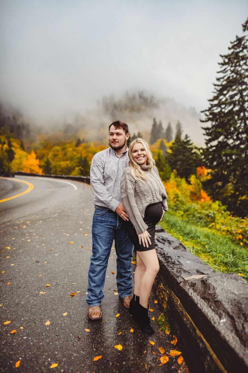 A couple laughs together on a foggy mountain road with autumn colors in the background.
