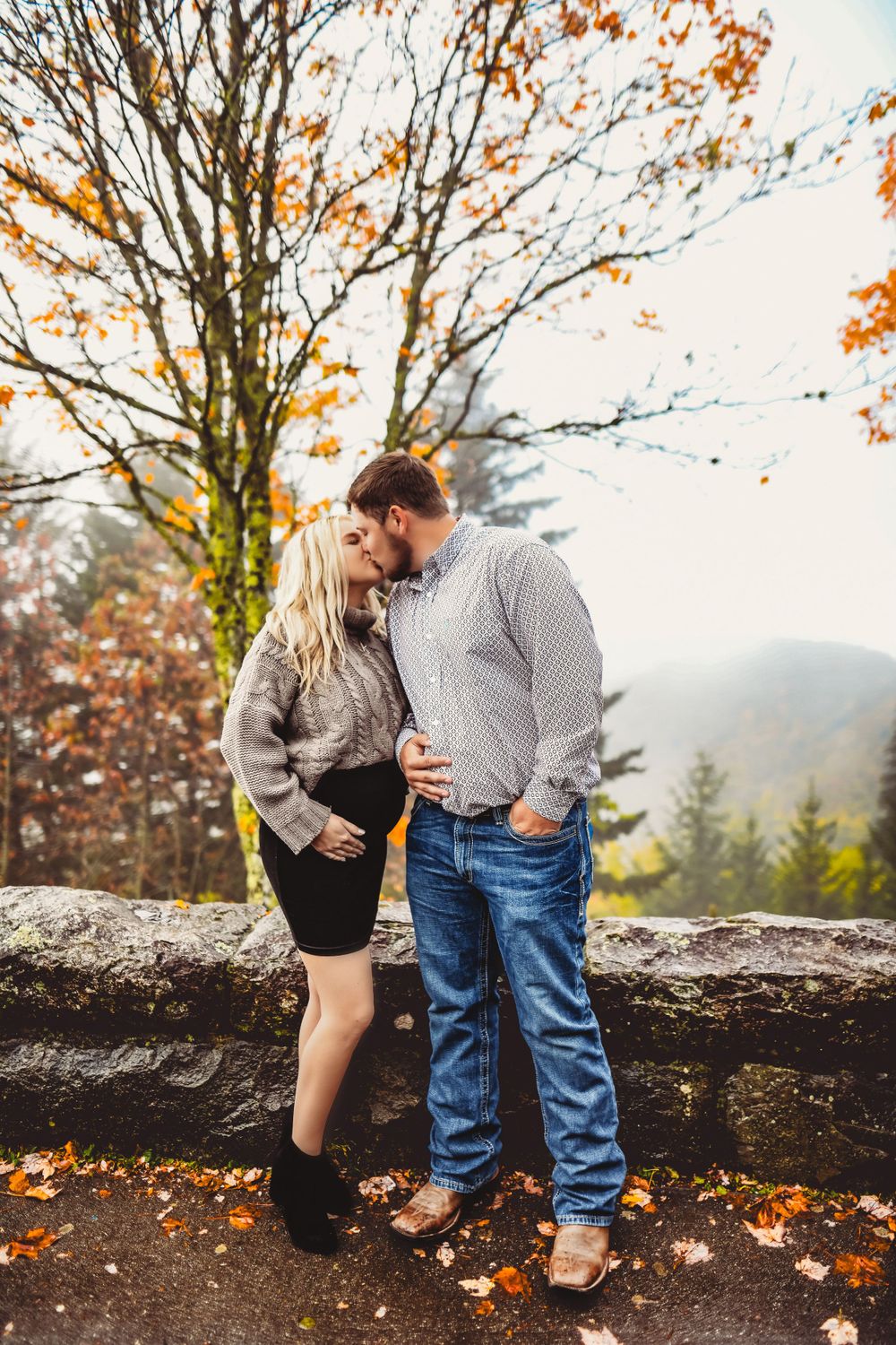 A couple shares an intimate moment on a stone wall surrounded by fall leaves.