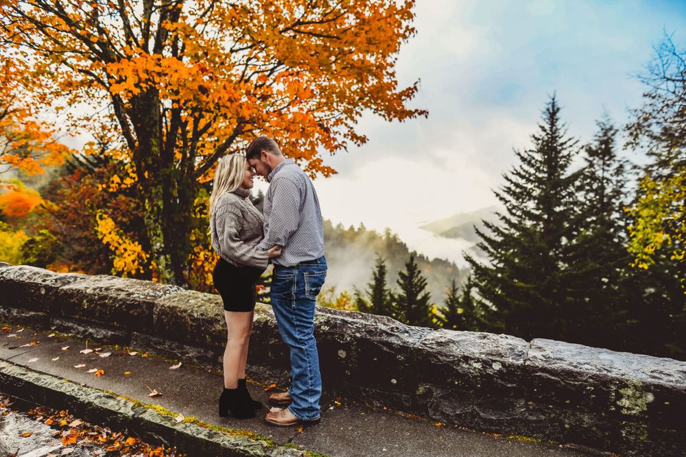 A couple stands together on a scenic overlook with autumn trees and mountains in view.