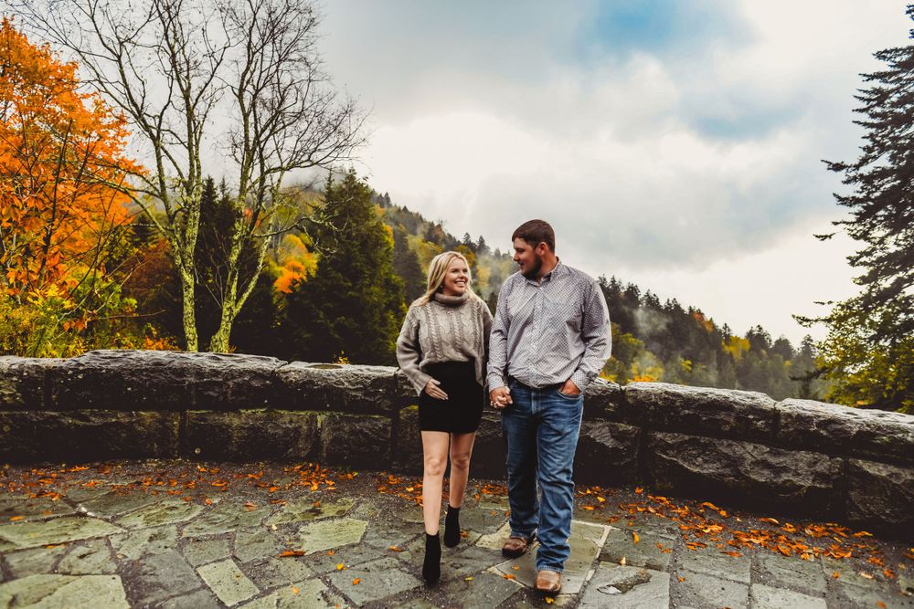 A couple enjoys a romantic moment on a stone wall with fall foliage and mountain views.