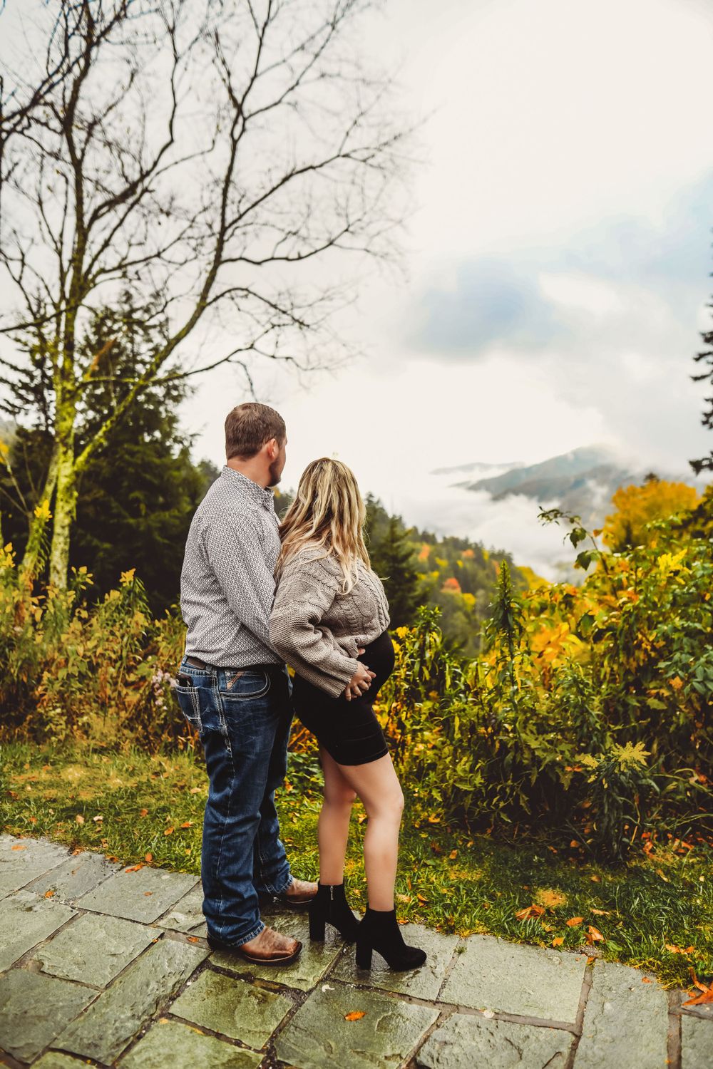 A couple stands close together on a stone path with misty mountains and autumn colors in the background.