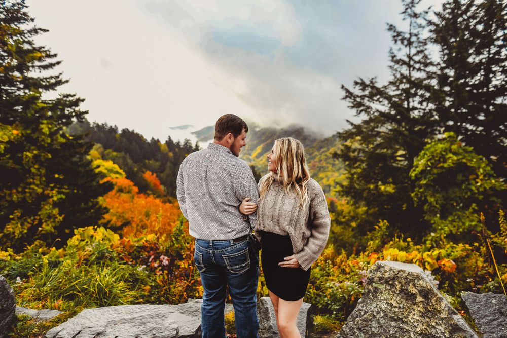 A couple poses on a mountain overlook surrounded by vibrant fall colors and fog.