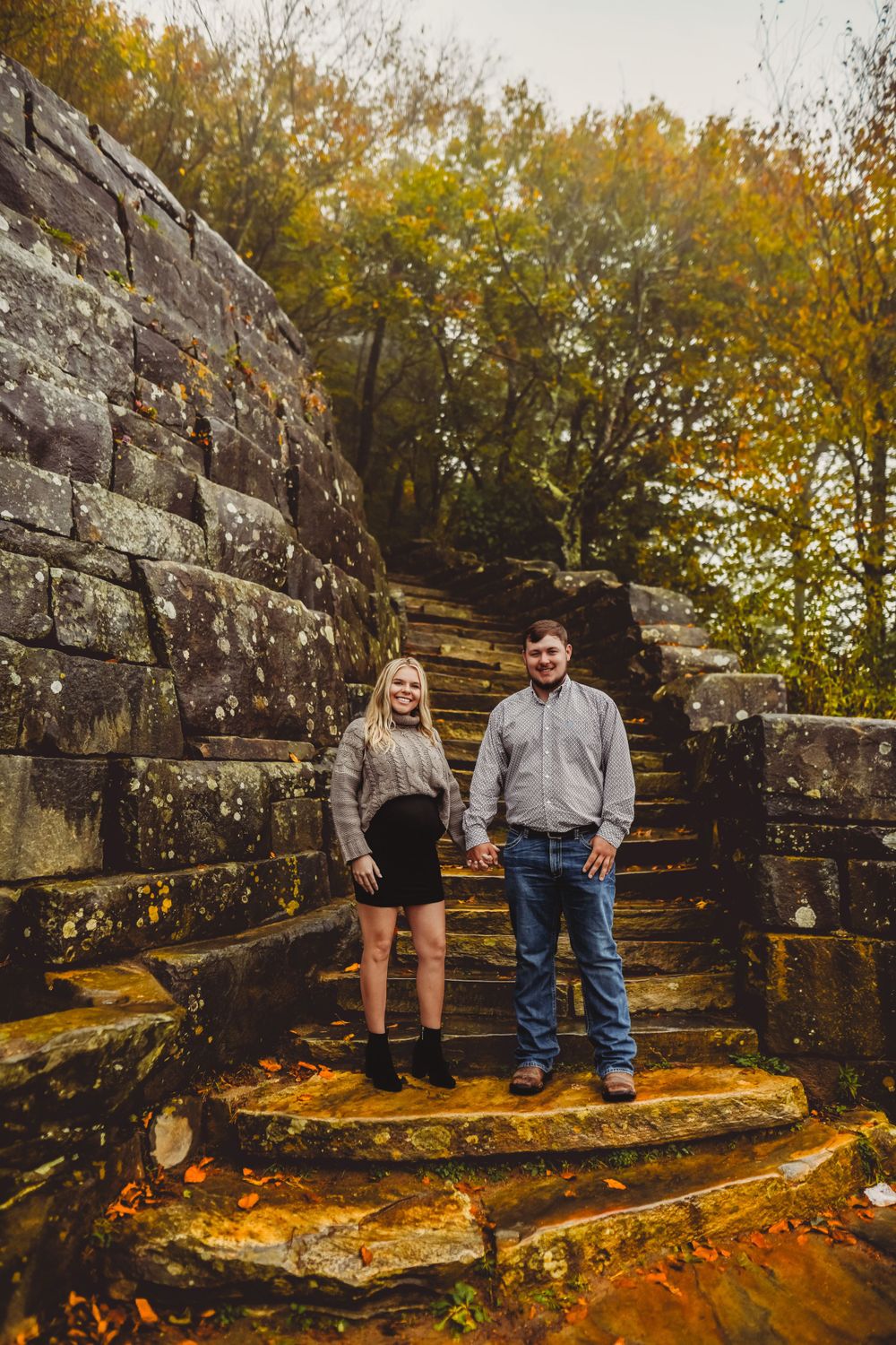 A couple stands on stone steps with a rustic stone wall and autumn foliage in the background.