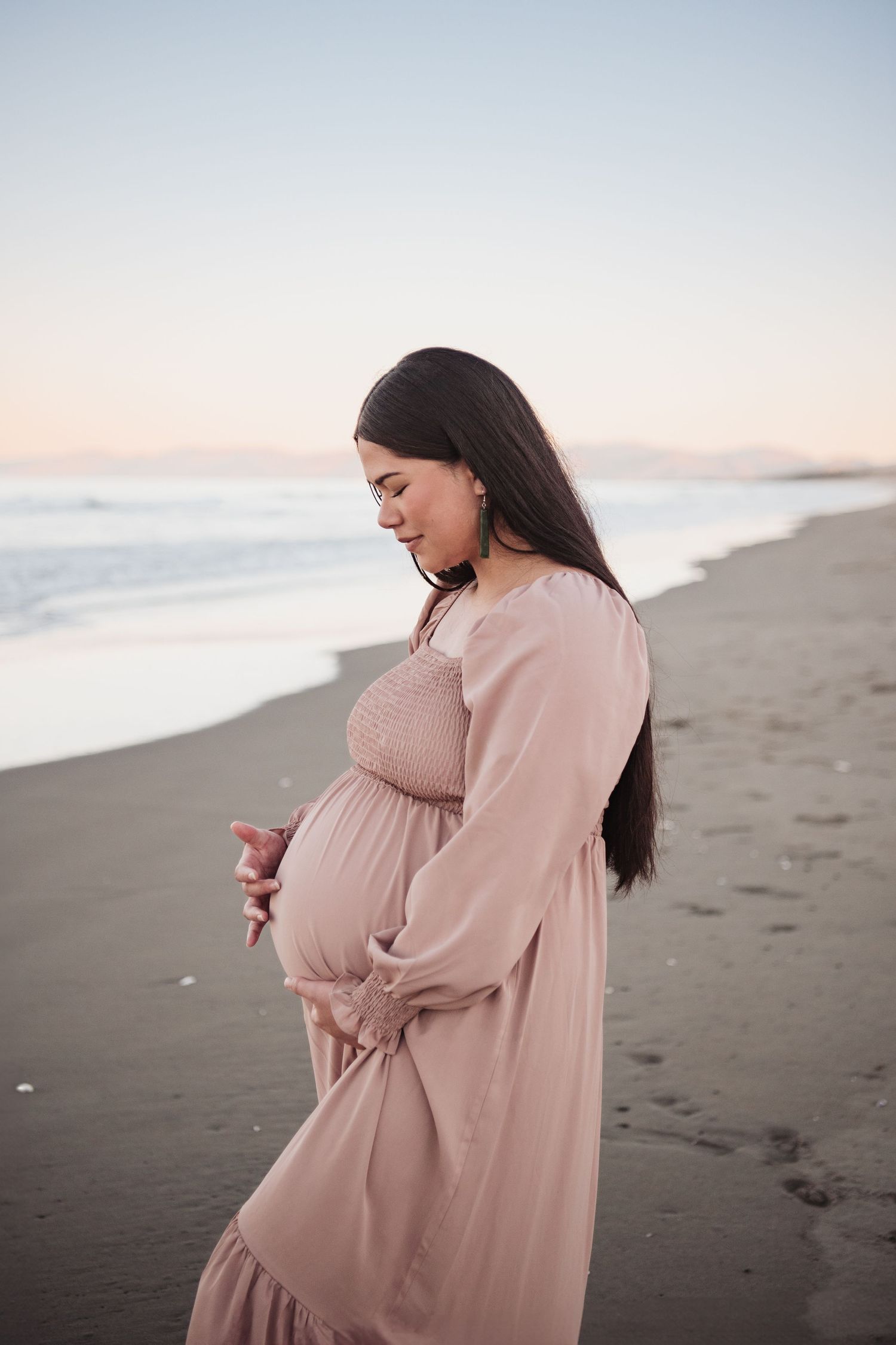 Expectant mother in pink flowy dress standing on beach at sunset.