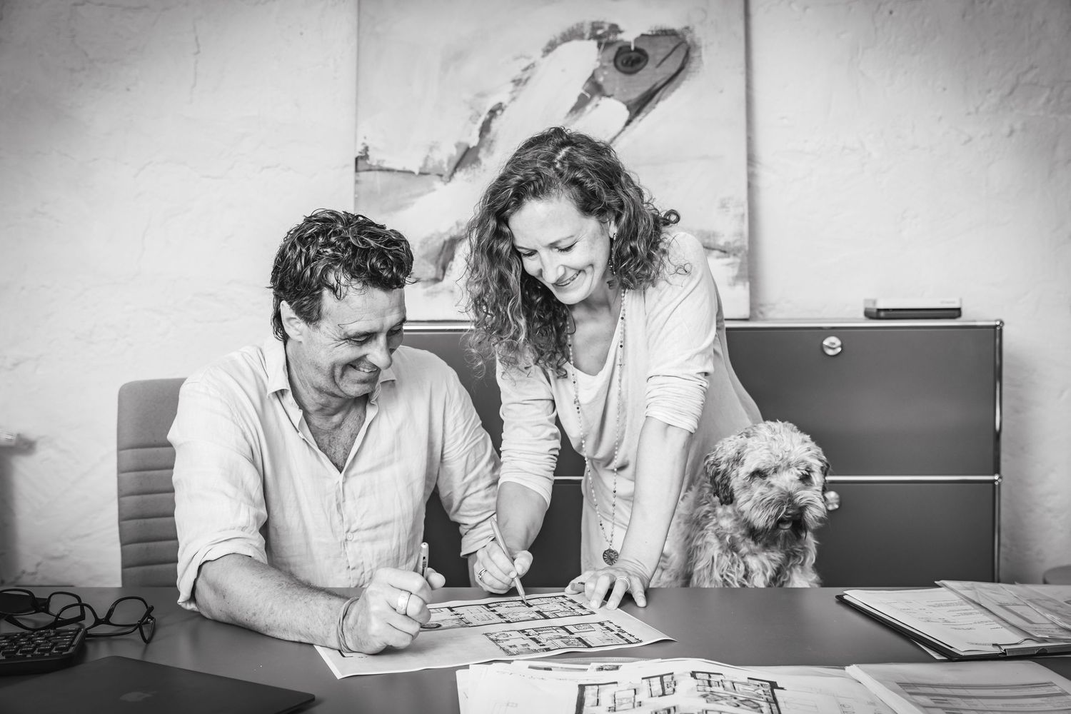 Black and white lifestyle photoshoot of a couple in an office at a table with a fluffy dog looking at photos, smiling.