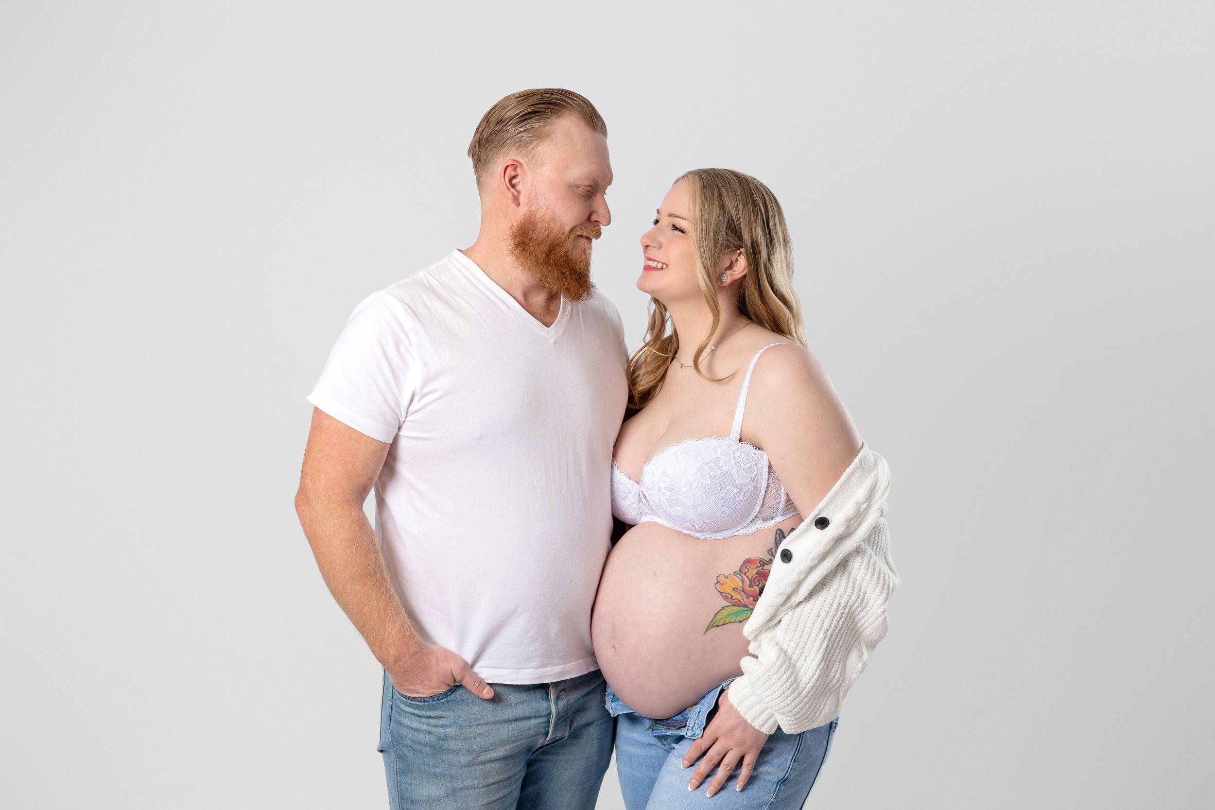 A couple in casual white attire share an intimate moment against a light gray studio backdrop.