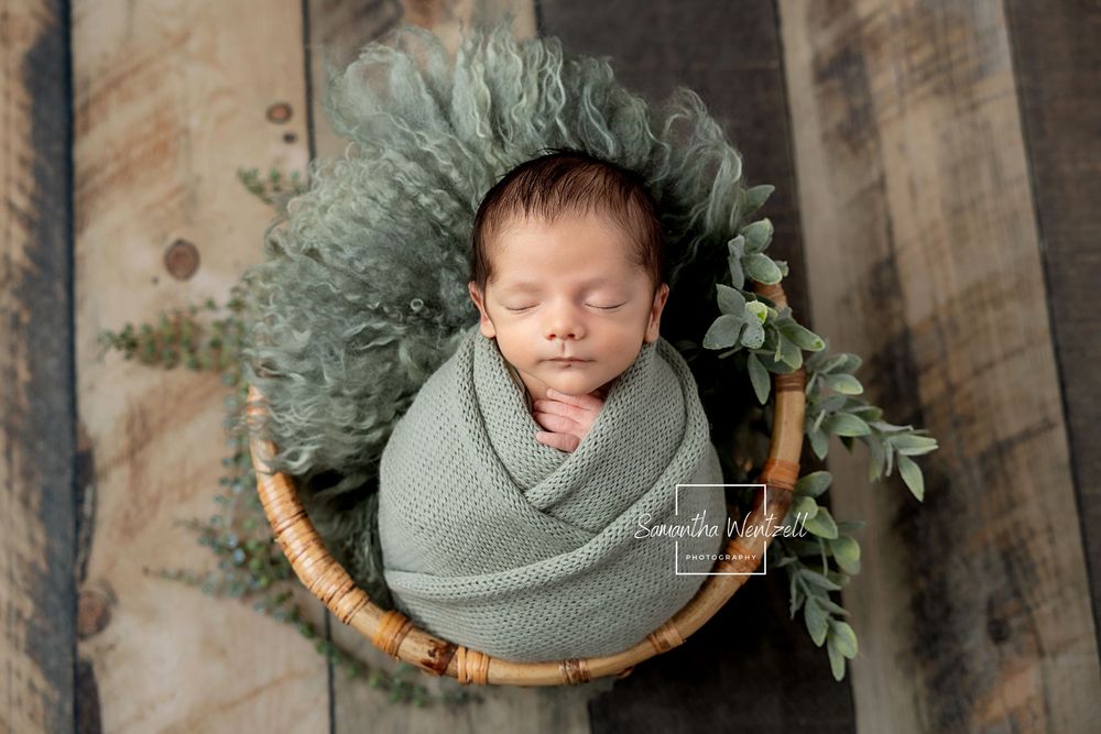 Newborn baby wrapped in green swaddle sleeping peacefully in a wicker basket with eucalyptus leaves.