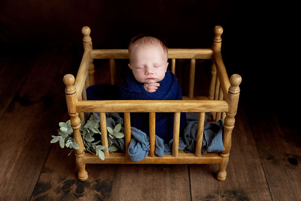 Newborn baby sleeping peacefully in a tiny wooden crib wrapped in a blue knit blanket with green leaf accents.