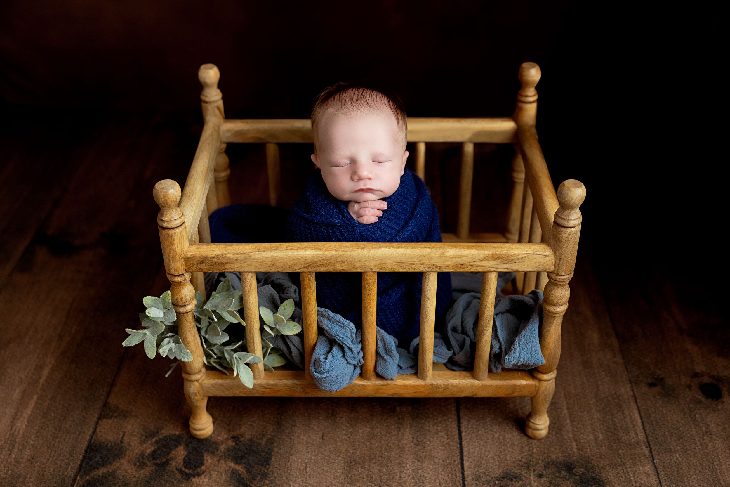 Newborn baby sleeping peacefully in a tiny wooden crib wrapped in a blue knit blanket with green leaf accents.
