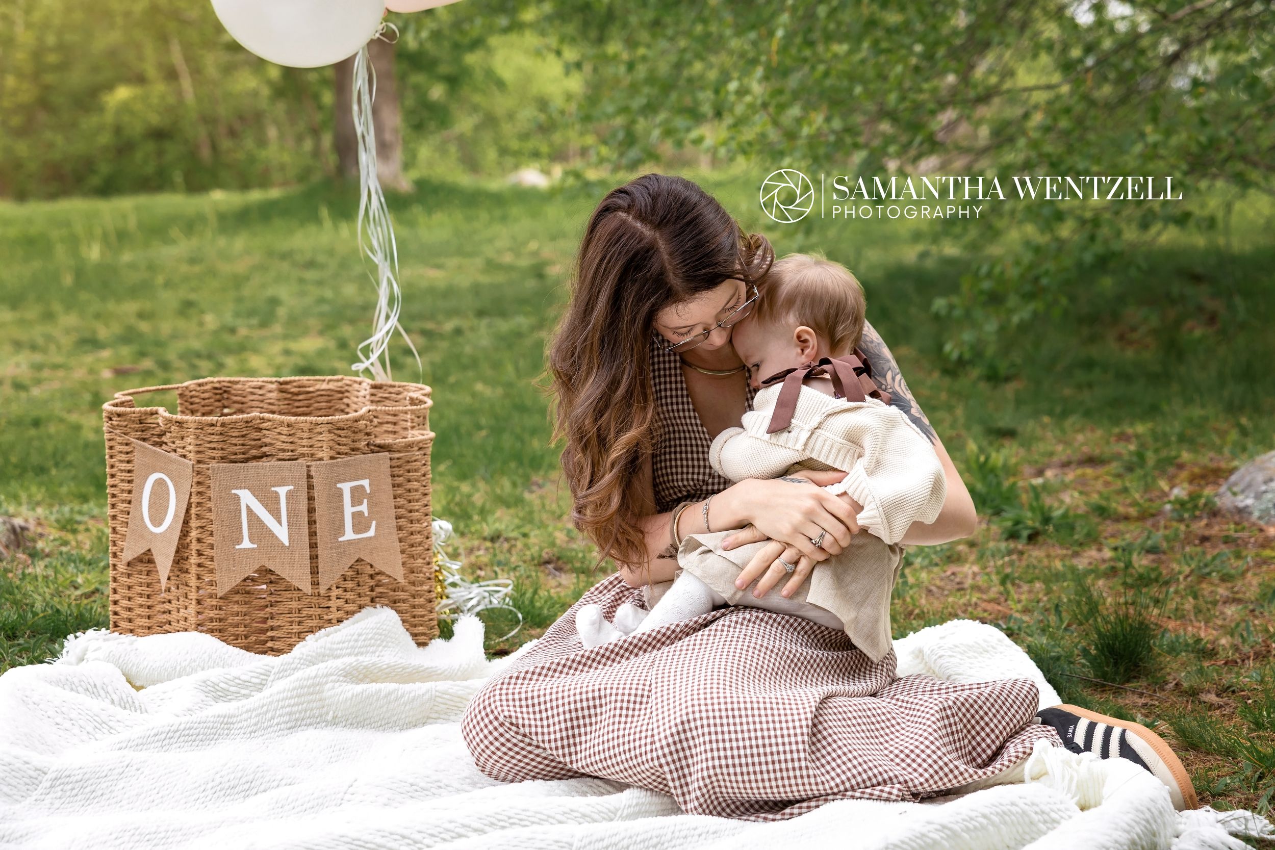 Mother kissing her baby during a first birthday outdoor photo session with ONE banner and balloon.