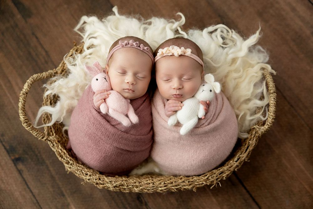 Two sleeping infants wrapped in soft pink swaddles lie together in a cozy woven basket.