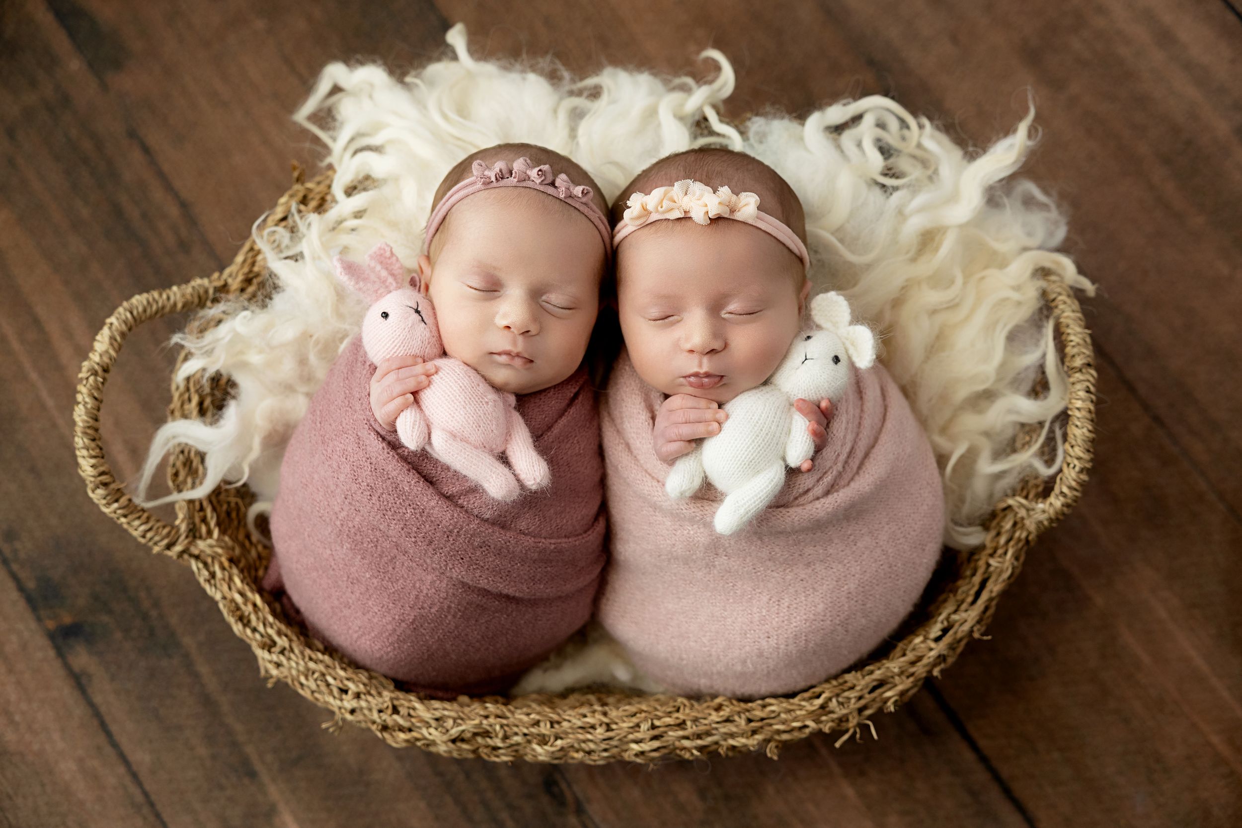 Two sleeping infants wrapped in soft pink swaddles lie together in a cozy woven basket.