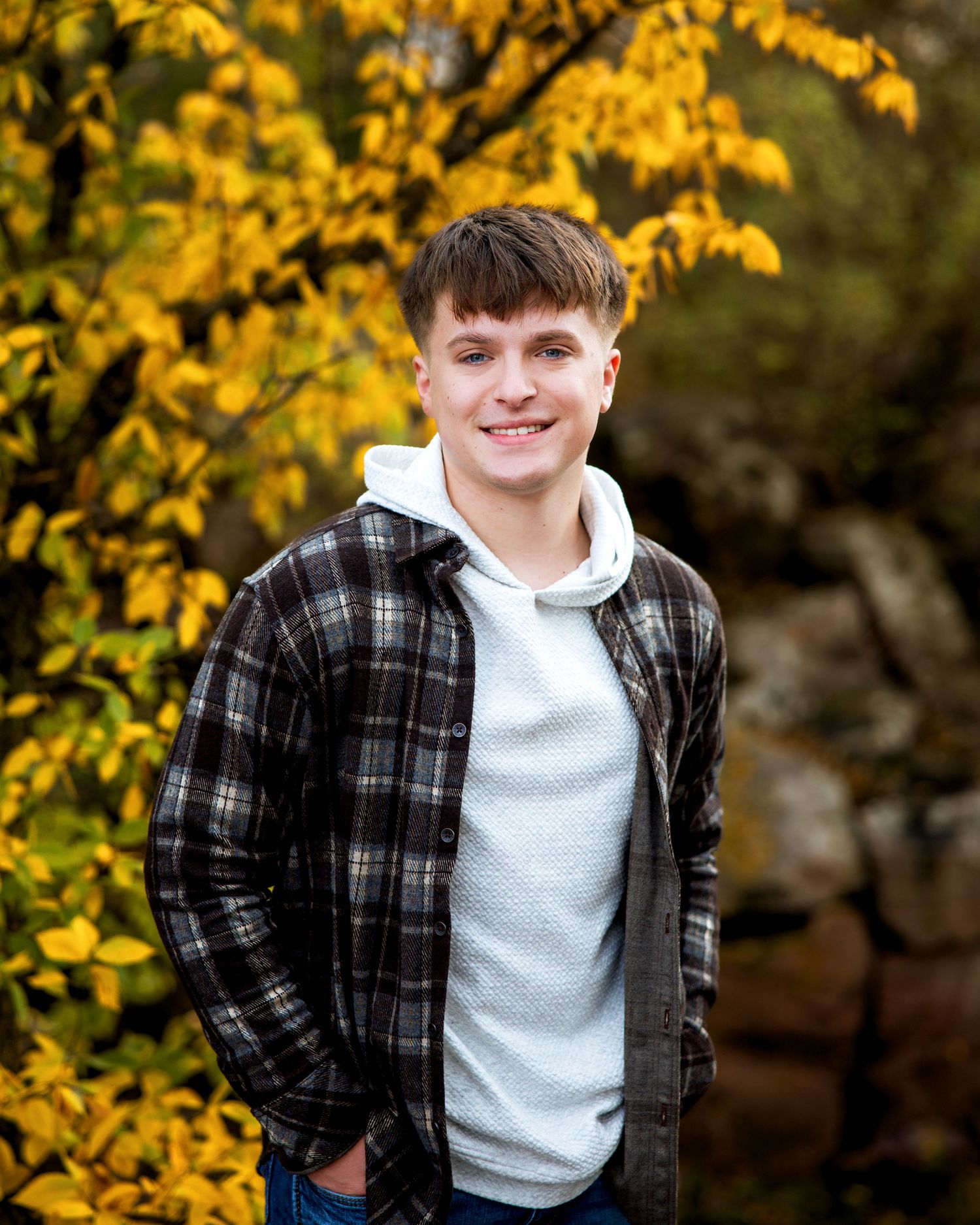 Smiling teen boy in plaid jacket and white hoodie poses outdoors among vibrant yellow autumn leaves.