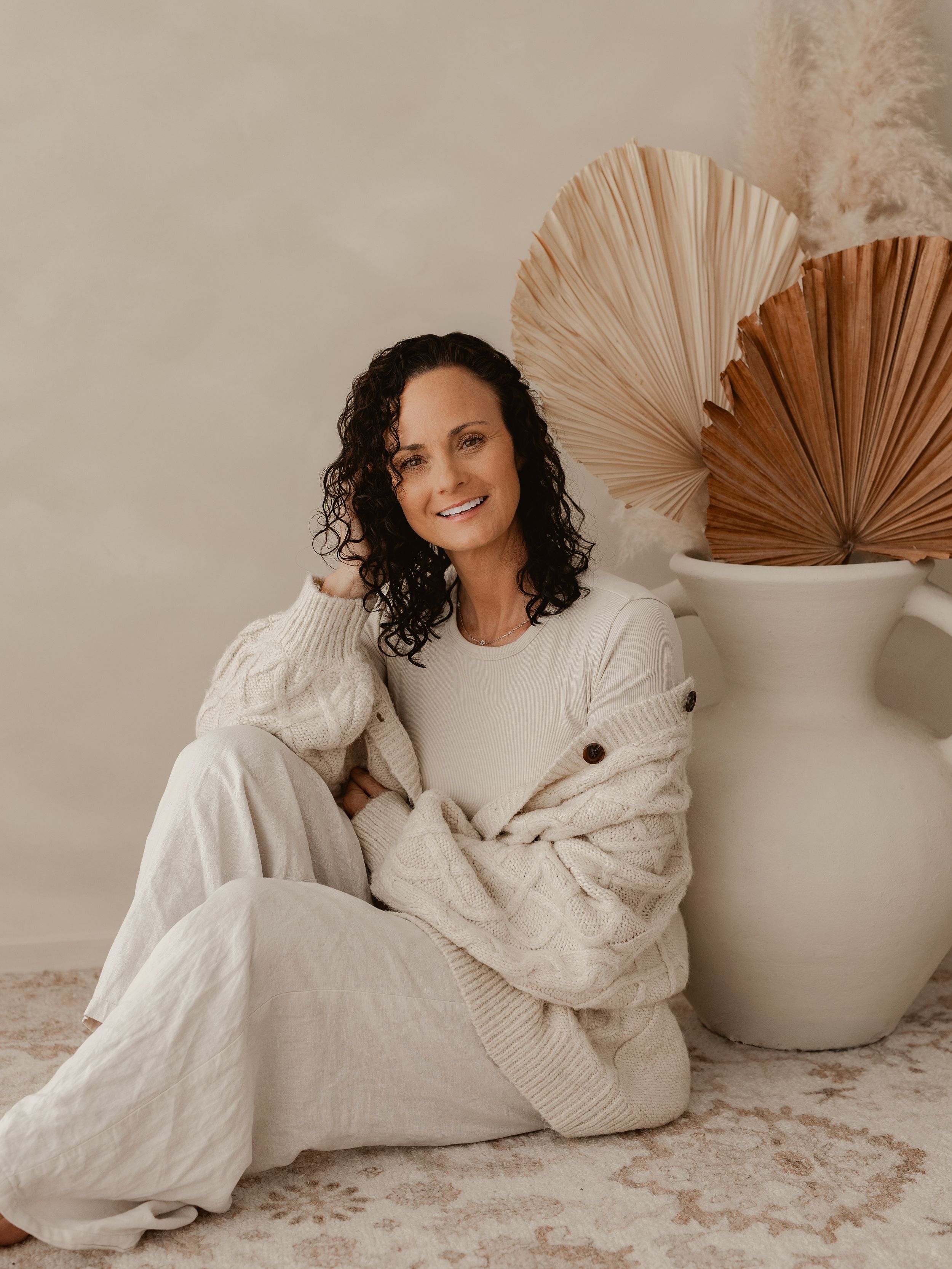 Person sits cross-legged wearing beige cable knit cardigan and white pants in cozy neutral room with dried palm decor.