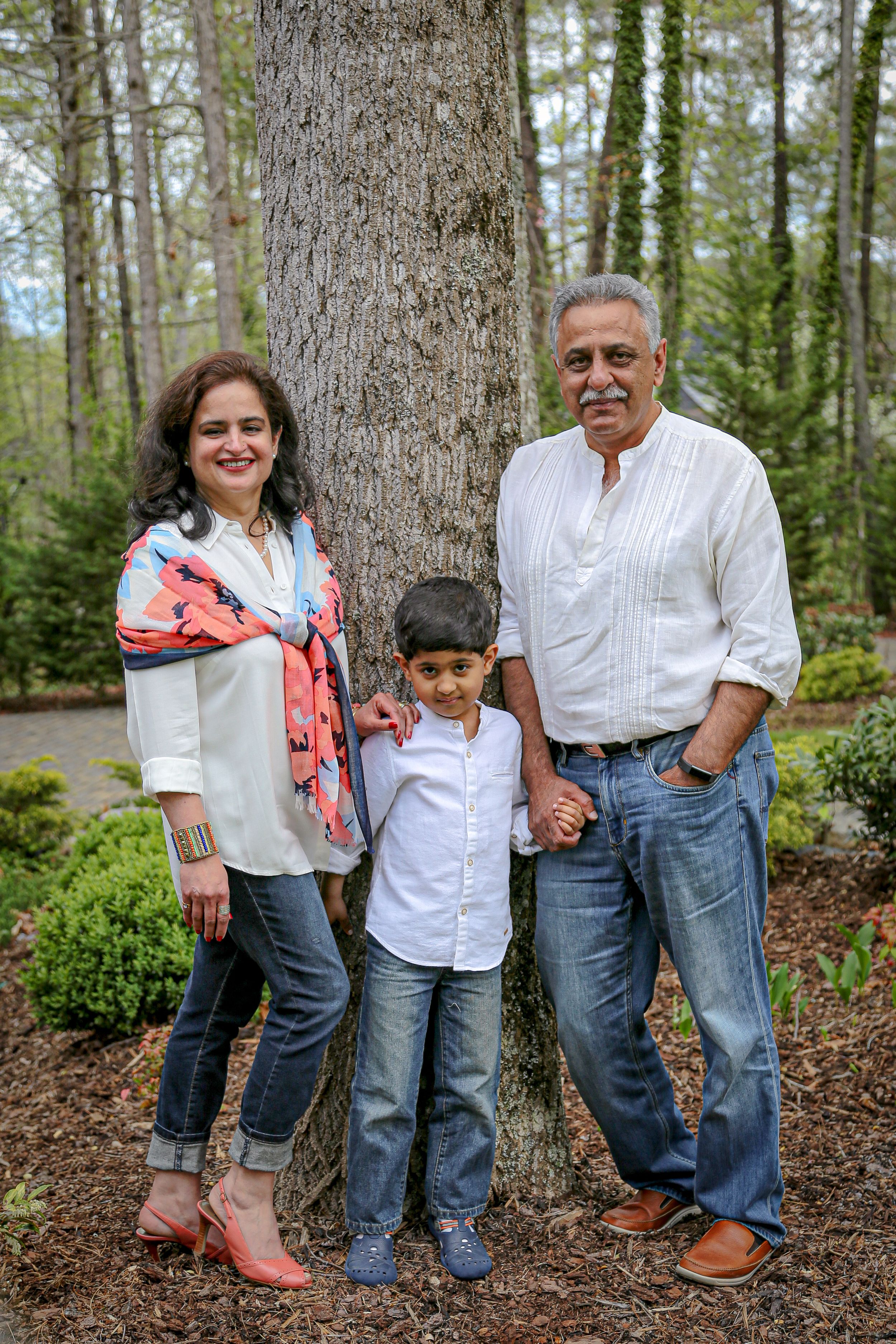 Three generations stand together in casual clothing against a tree in an outdoor setting.