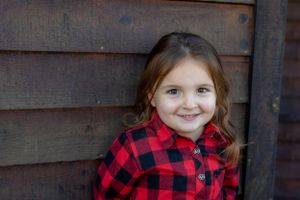 Child wearing red buffalo plaid flannel smiles against wooden wall.