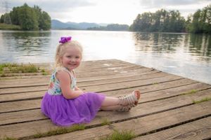 Young child in purple dress sits on wooden dock by serene lake at sunset.