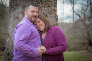 Couple in matching purple sweaters embrace by large tree trunk.