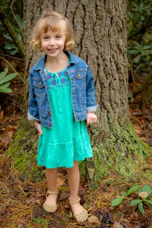 Young child in a turquoise dress and denim jacket poses happily in front of a large tree trunk.