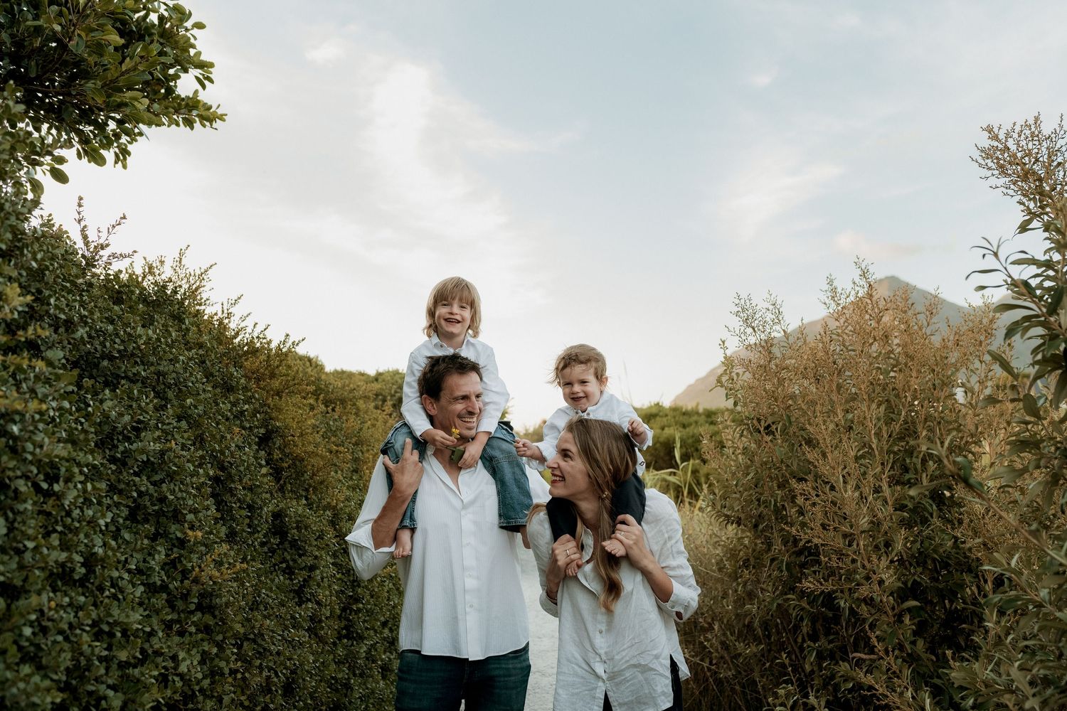 Happy family of four walking outdoors, parents carrying children on shoulders, surrounded by lush green hedges.