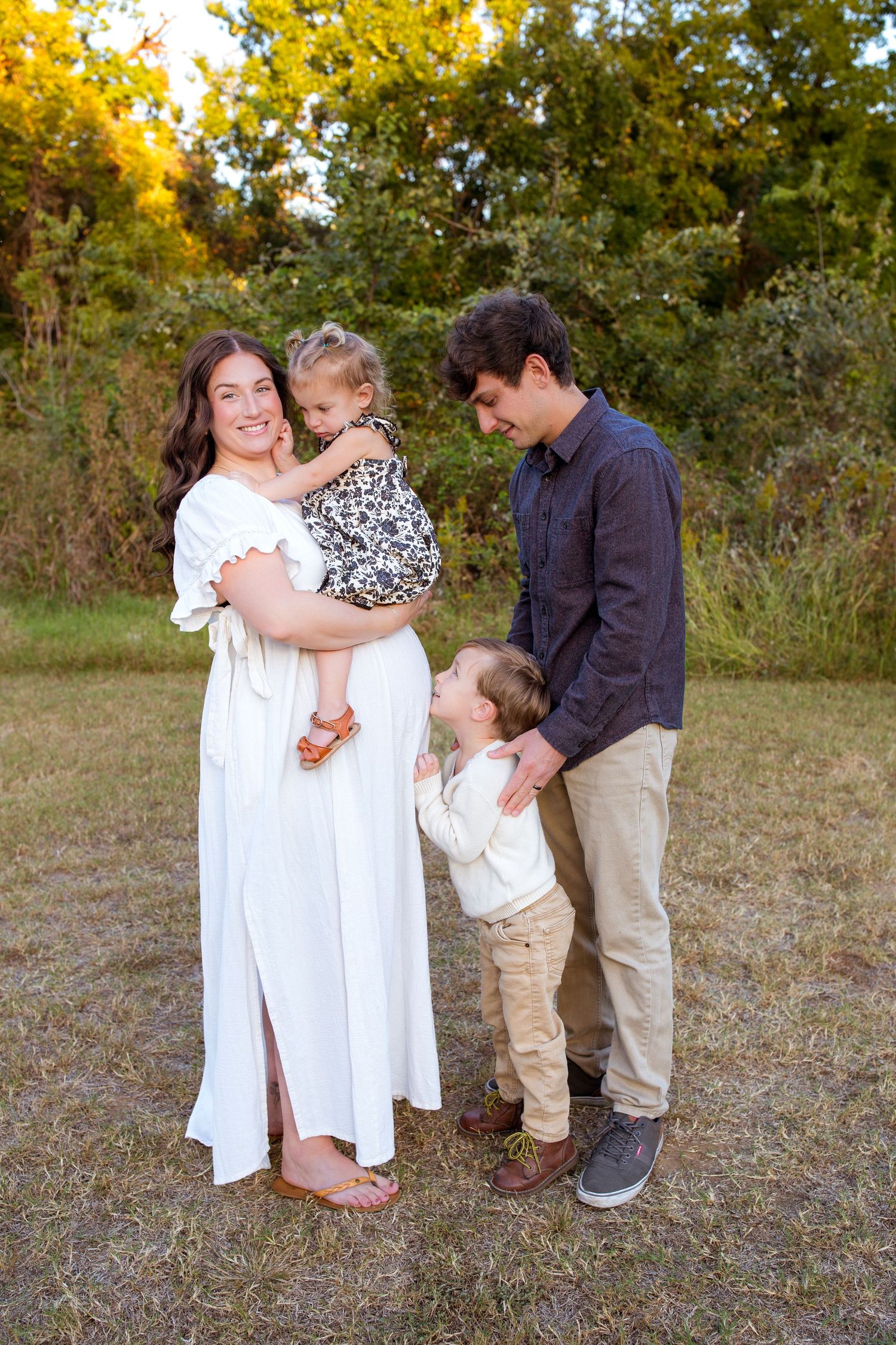 Pregnant woman in white dress with husband and two young children in an outdoor field.