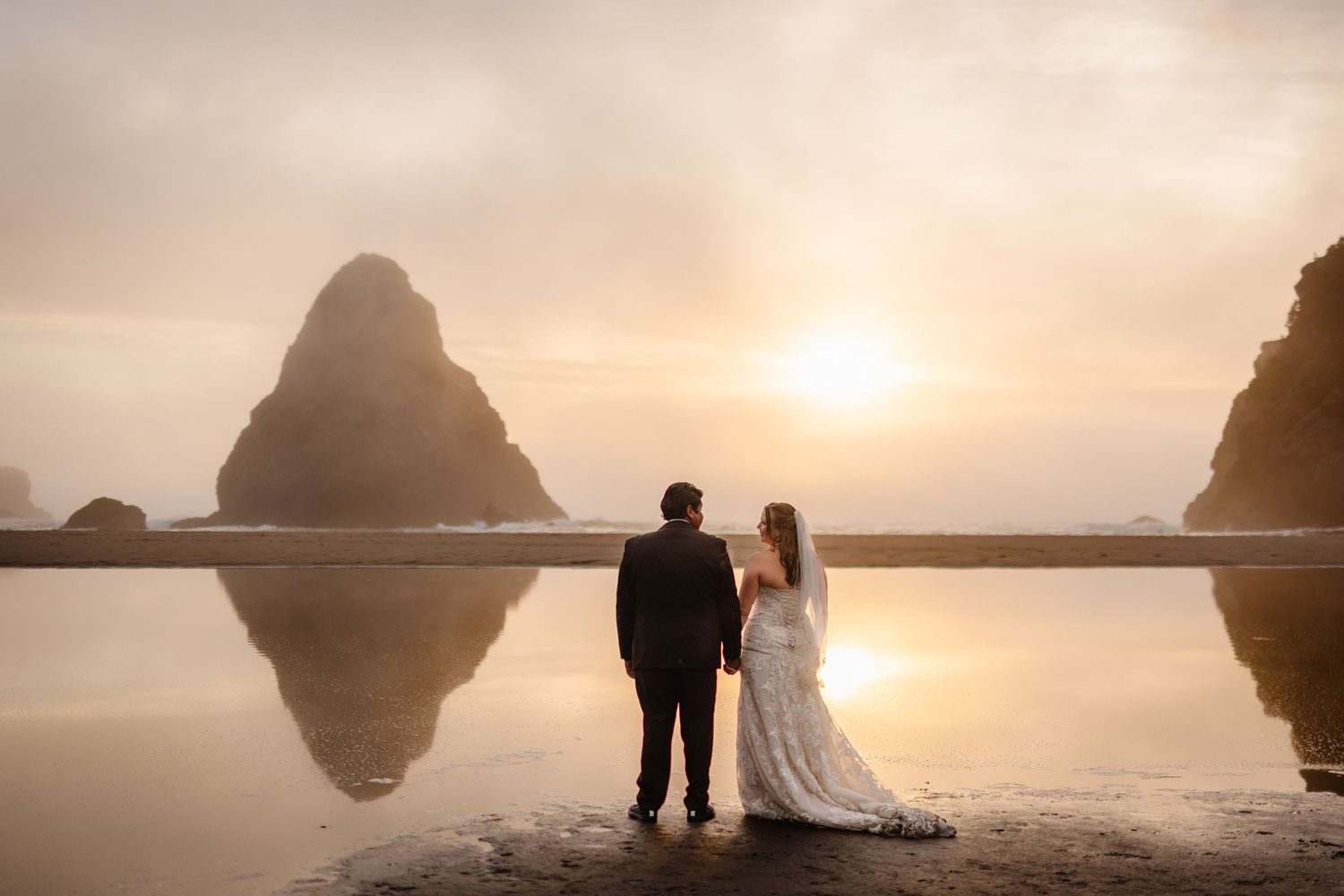Newlyweds hold hands while gazing at a stunning golden sunset reflected on the beach with towering sea stacks.