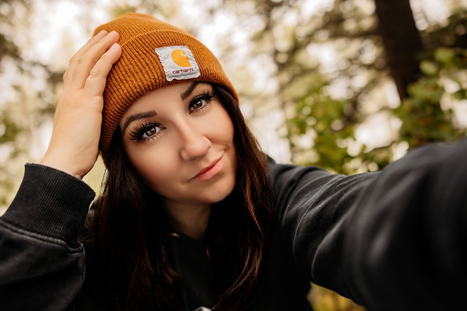 Someone wearing a brown Carhartt beanie and black sweater poses outdoors with autumn foliage in the background.