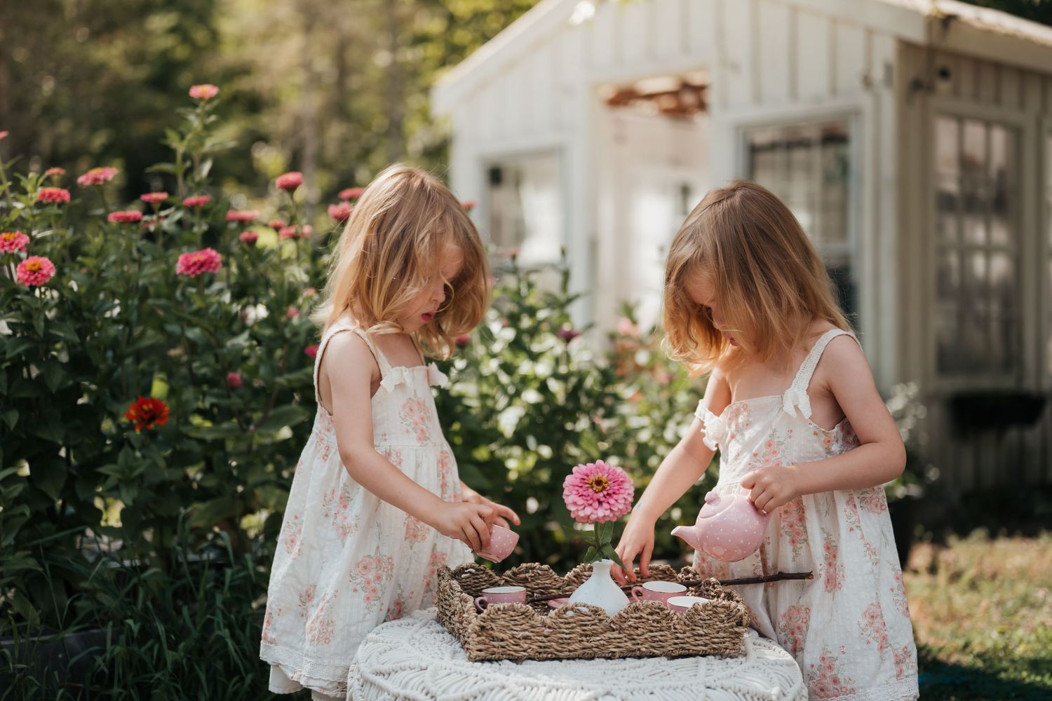 Two young girls in floral dresses picking flowers from basket in garden.