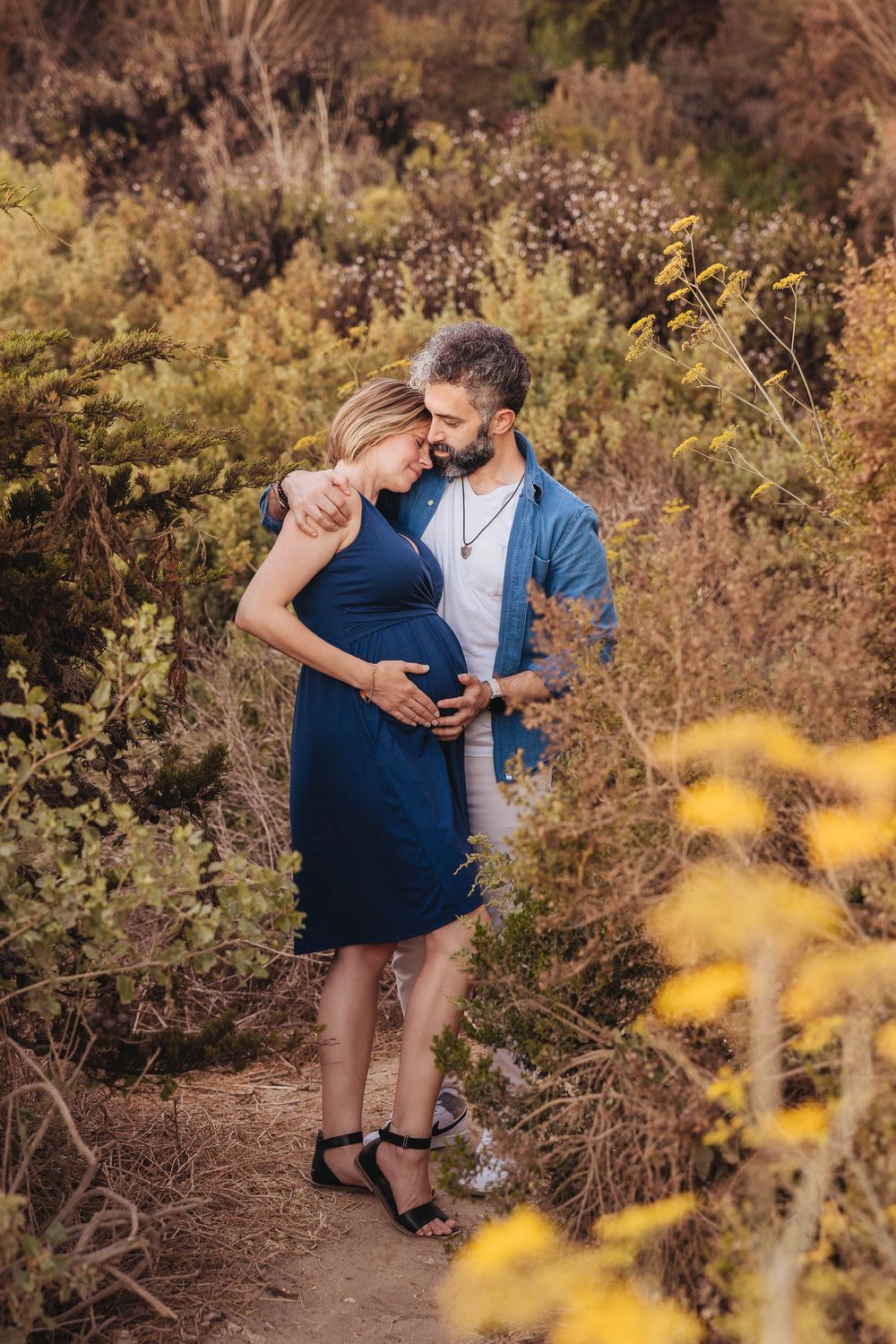 A romantic maternity photo of a couple embracing on a nature trail surrounded by golden wildflowers.