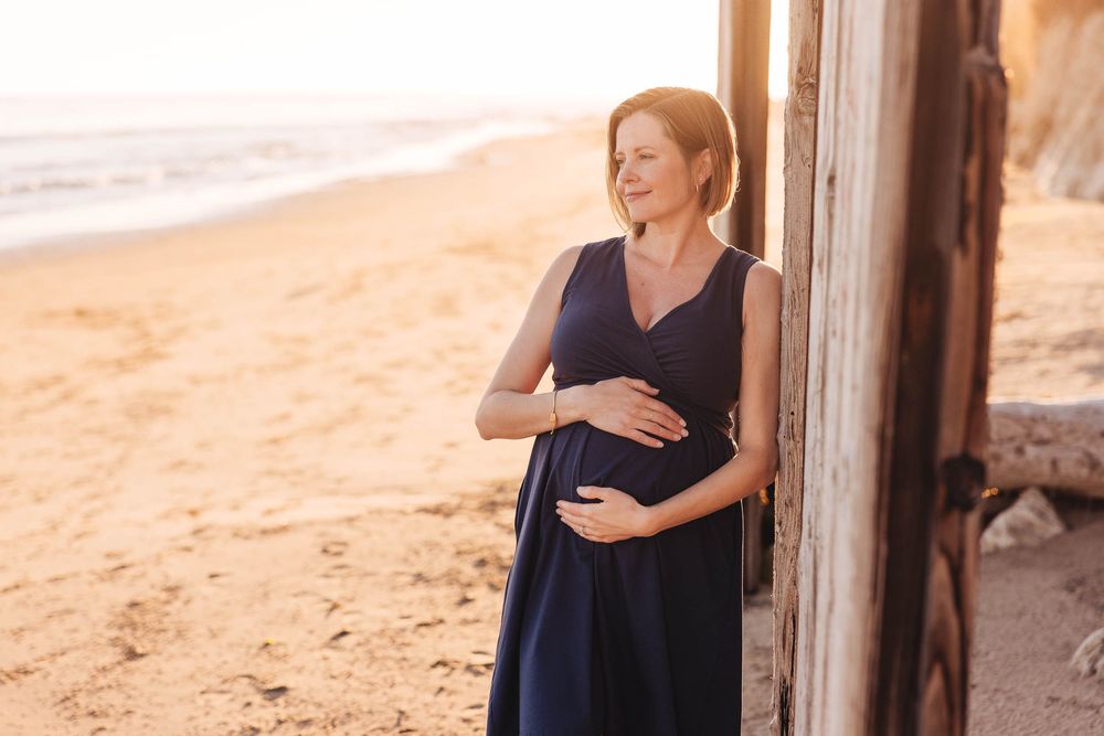A woman in a navy dress stands barefoot on a sandy beach during golden hour.