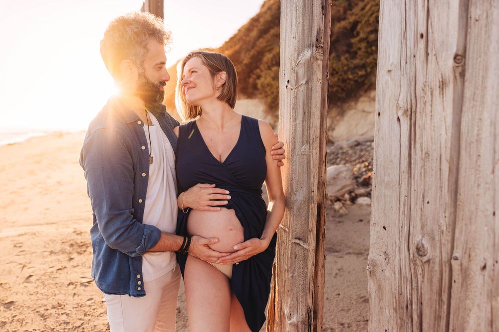 Romantic beach maternity photos captured between wooden posts at sunset.