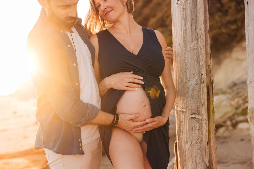 Tender maternity moment photographed against wooden pier posts during sunset.