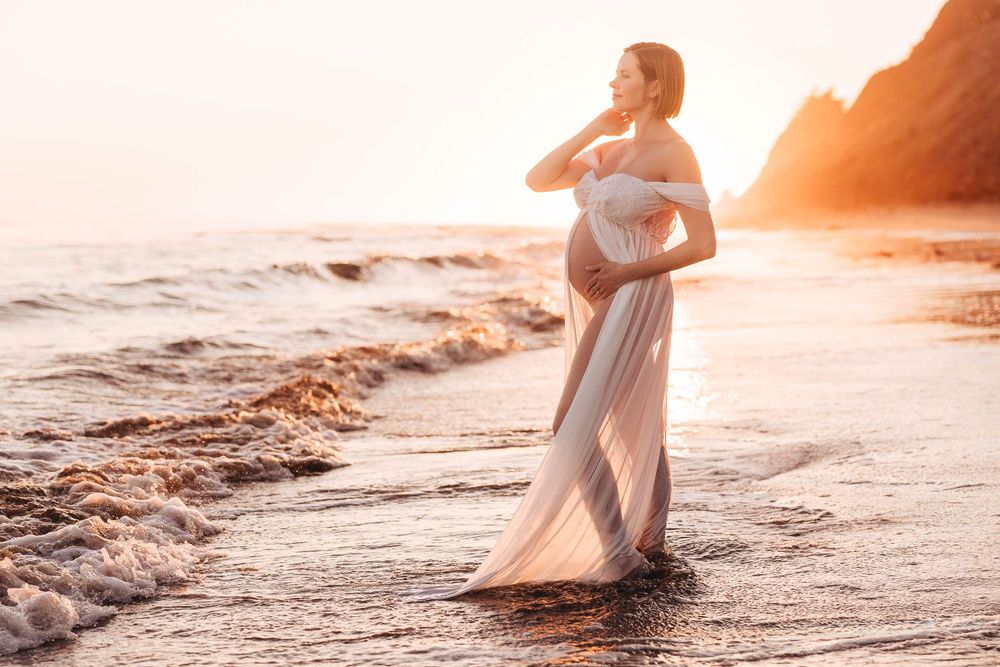 Woman in flowing white dress walks along shoreline during sunset with cliffs in background.
