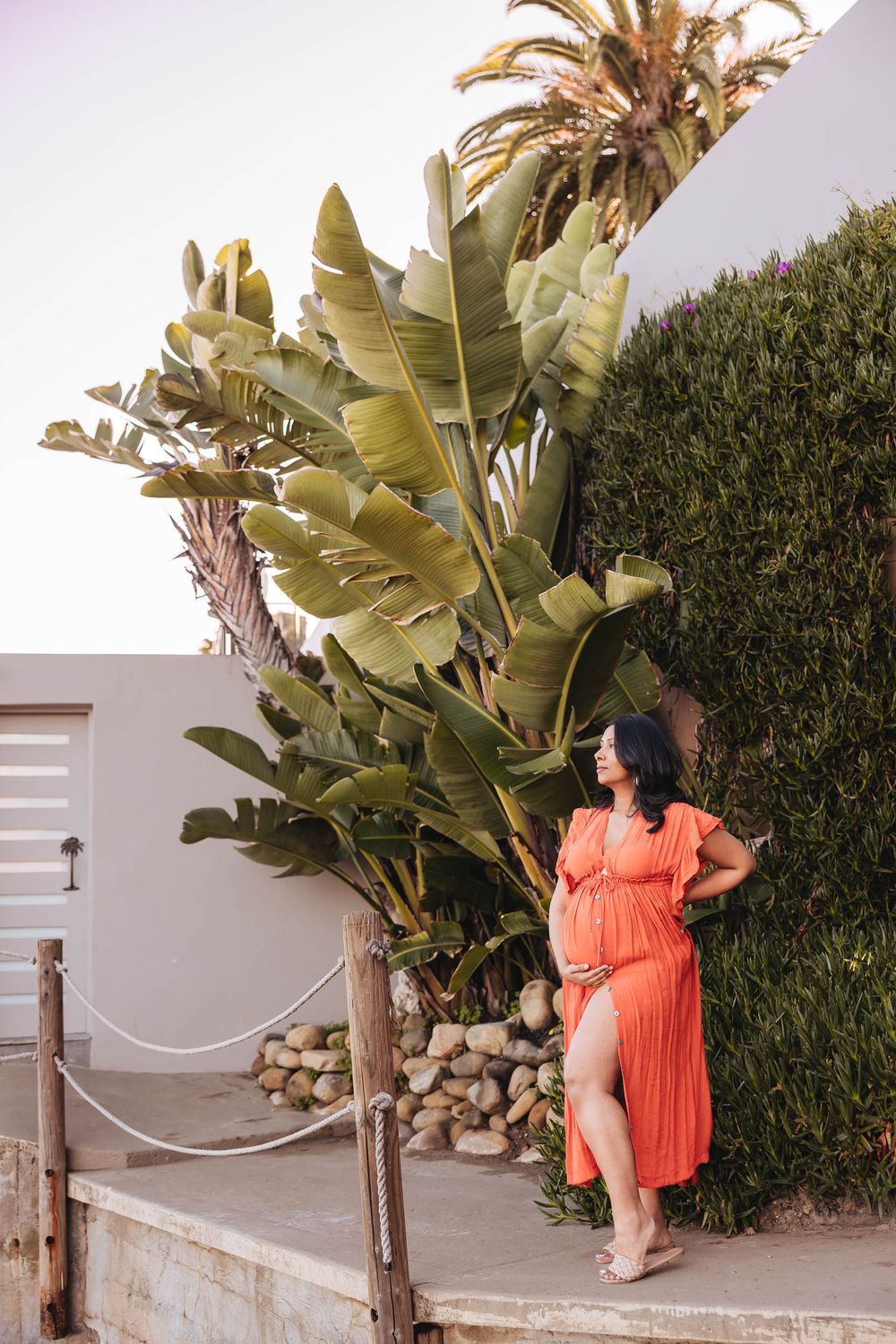 A woman in a coral dress poses against a white wall with tropical plants and greenery in an outdoor setting.