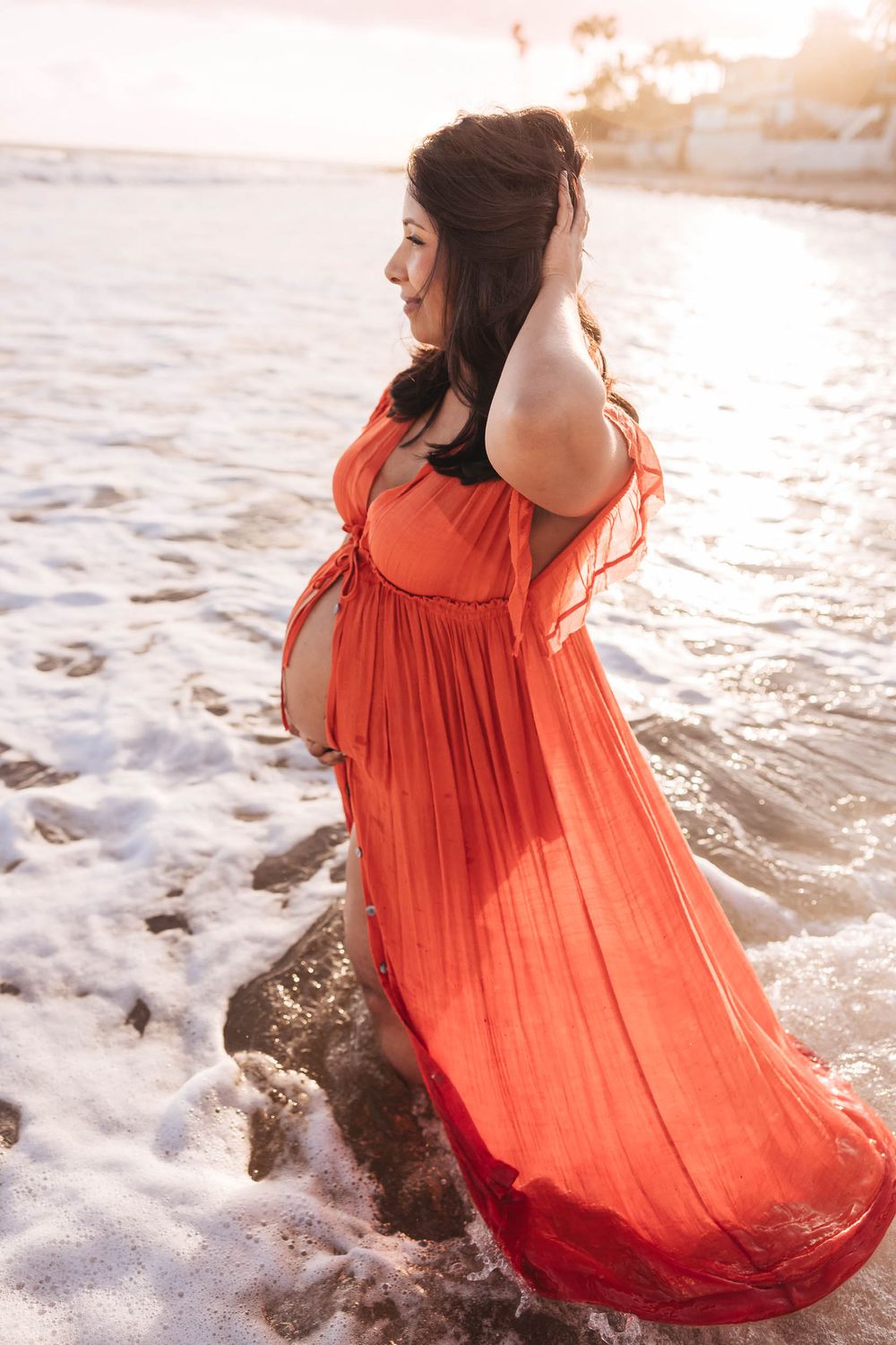 Woman in flowing coral dress walking along beach shoreline at sunset as waves crash against rocks.