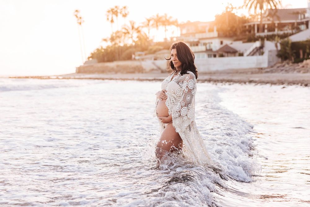 A person in a white lace dress stands in shallow ocean waves at sunset on a tropical beach.