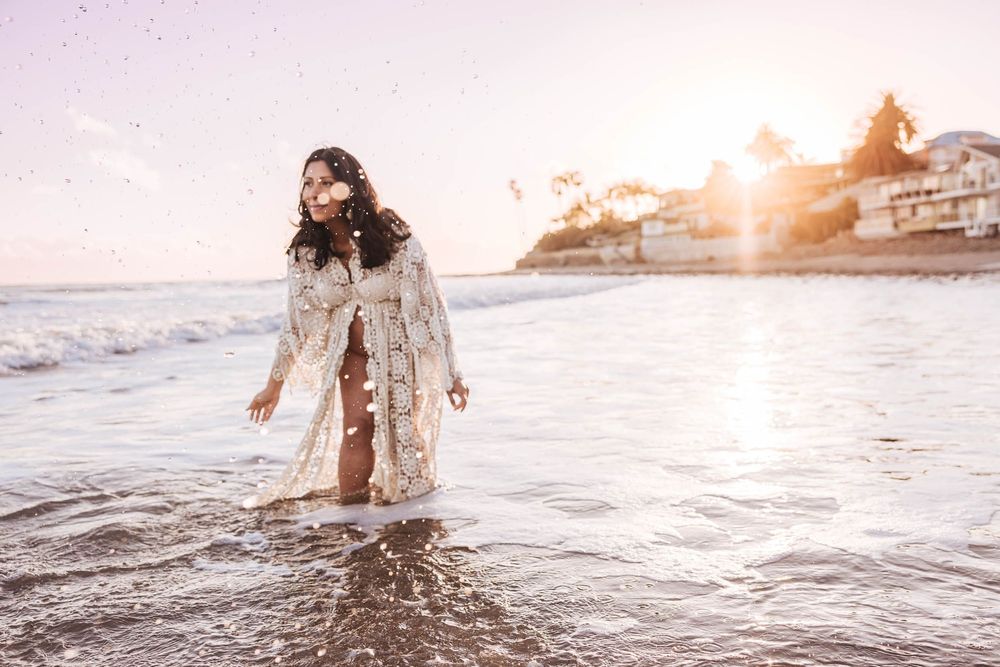 A person in a white lace dress stands in shallow ocean waves at sunset with coastal homes in the background.