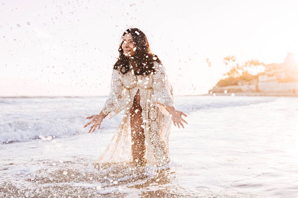 Person in flowing white dress splashing in ocean waves at sunset with golden light creating dreamy atmosphere.