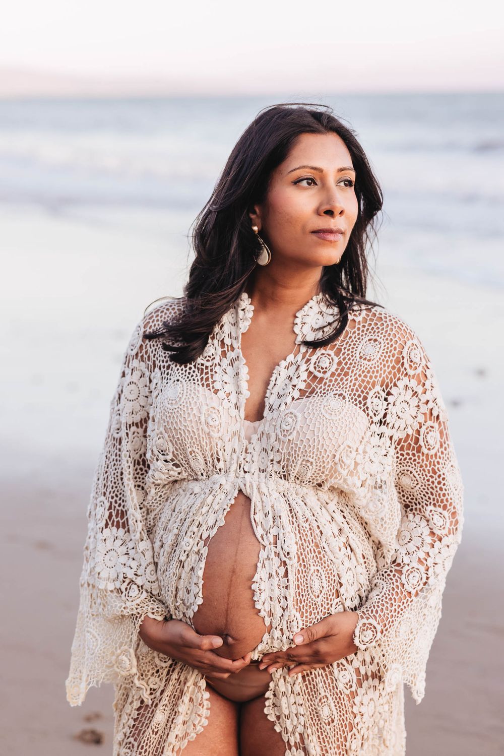 A woman in a delicate white lace shawl poses gracefully on a beach at sunset.
