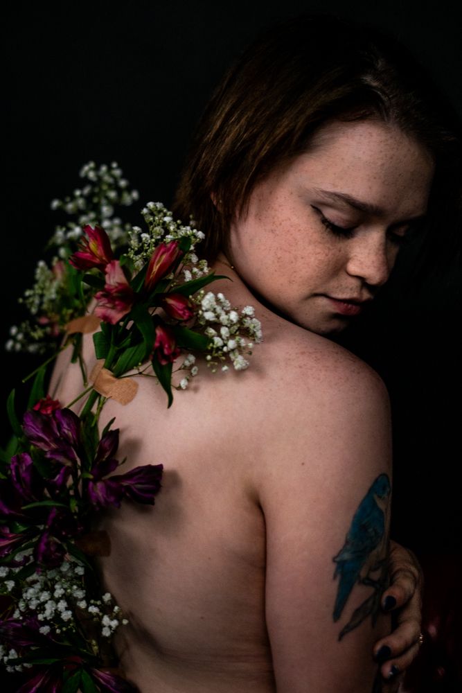 Artistic portrait featuring flowers and baby's breath arranged on bare shoulders with a visible tattoo in moody lighting.