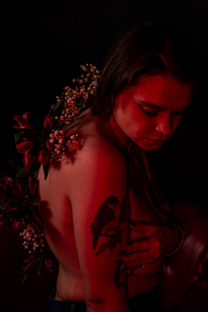 Artistic portrait in red lighting showing a figure adorned with berries and flowers in a moody, dark setting.