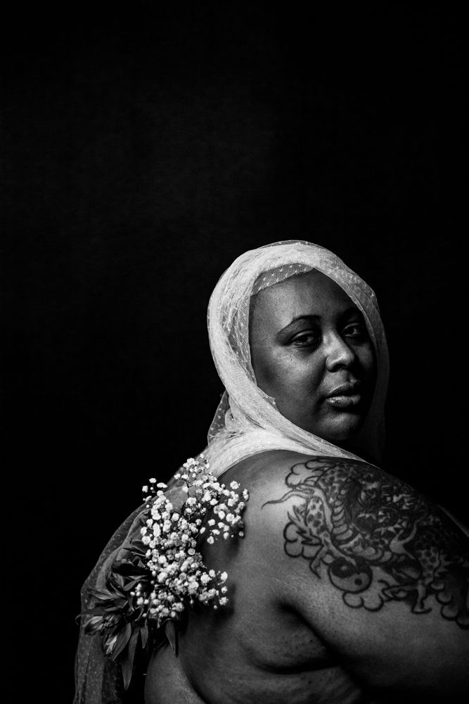 Artistic black and white portrait showing a draped figure with shoulder tattoos and baby's breath flowers against dark background.