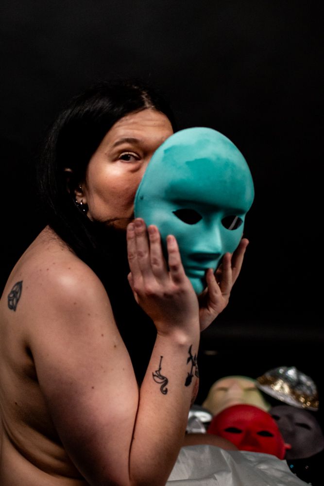 A dramatic portrait showing someone holding a turquoise theatrical mask against a black background.