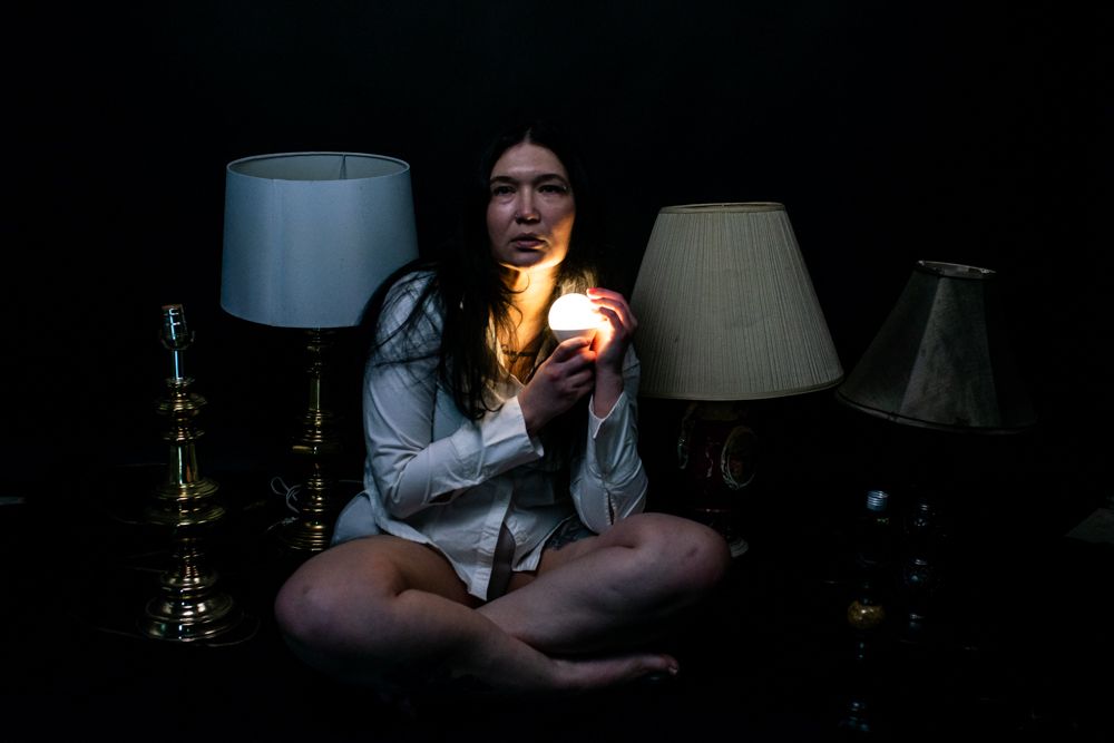 A moody dark photo with someone sitting cross-legged between table lamps holding a glowing light.