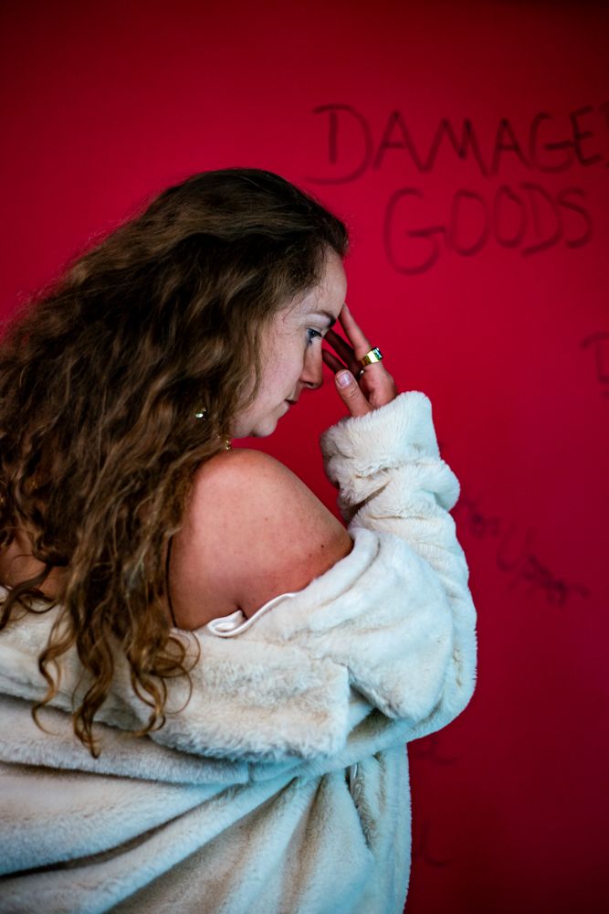 Side profile portrait against red wall with text showing person in white sweater with long wavy hair.