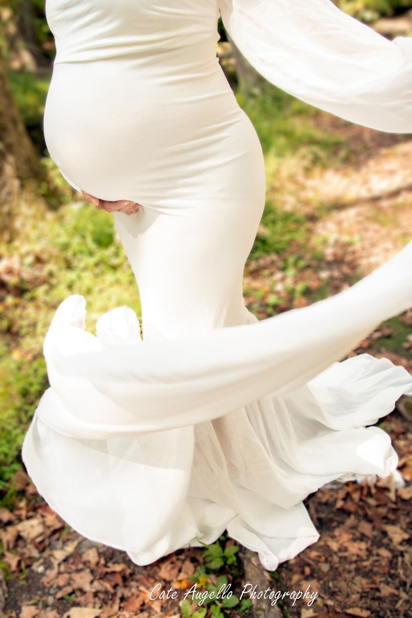 A flowing white maternity gown with a mermaid silhouette swirls elegantly against a natural outdoor background. Vernon, CT