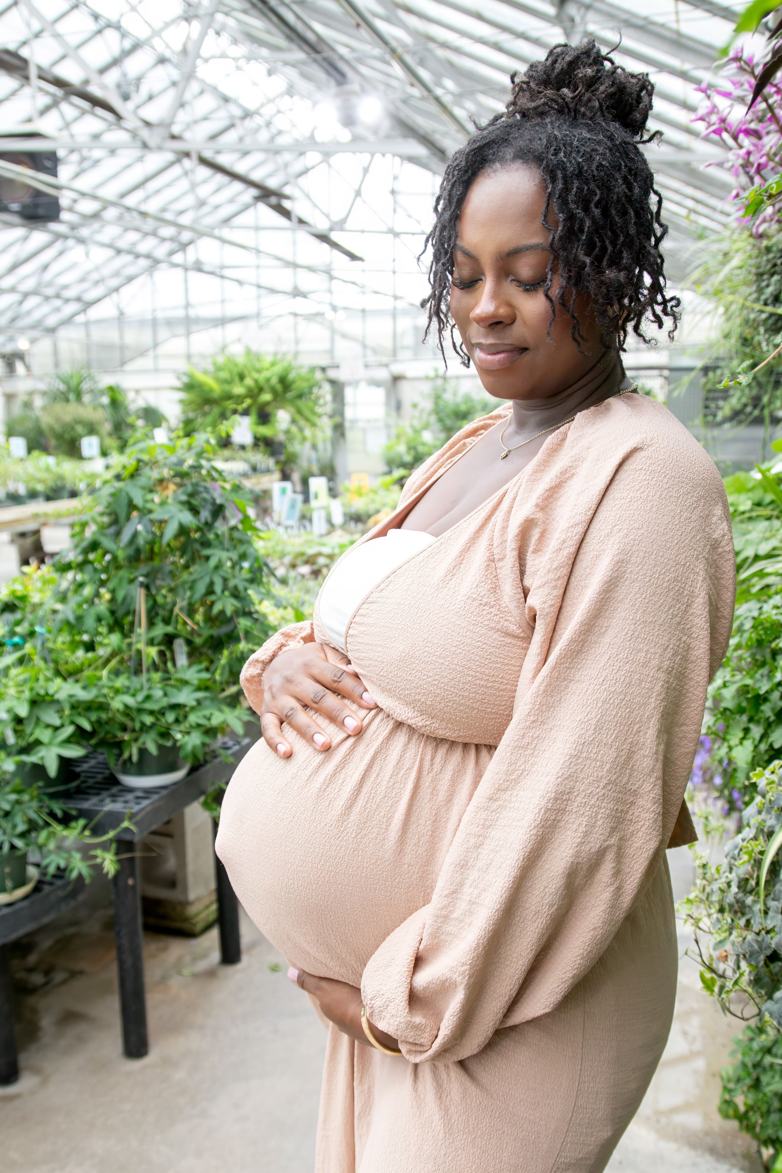 Pregnant Black woman in Hartford, Connecticut cradling her baby bump in a lush greenhouse filled with green plants. Maternity Photo session