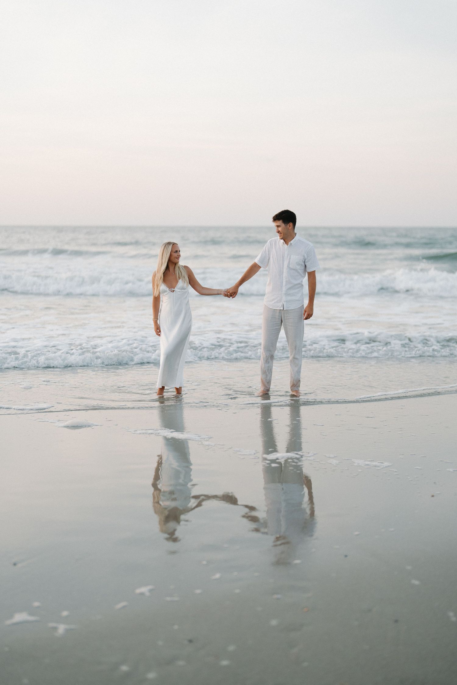 Couple in white attire holding hands on a serene beach at sunset with ocean waves behind them.