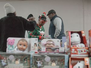Shoppers browse holiday merchandise at a counter displaying dolls and toys at a Christmas market.