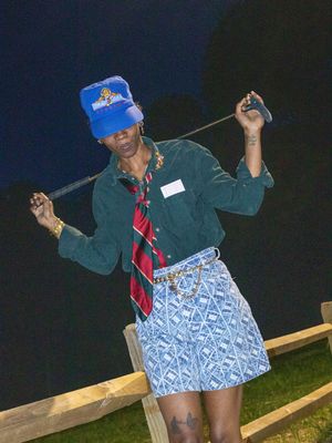 A person wearing a blue baseball cap, striped tie and dark green jacket poses at night with golf clubs.